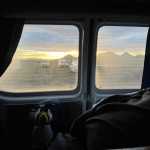 View from inside a campervan showing a sunset over distant Icelandic mountains through the window, with plush toys and a cozy blanket inside.