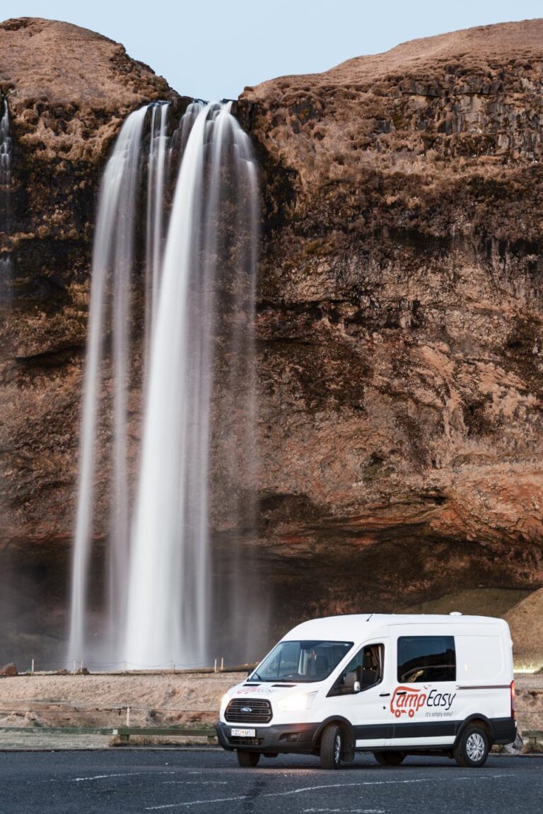A camping van parked in front of a waterfall surrounded by raw Icelandic rock formations.