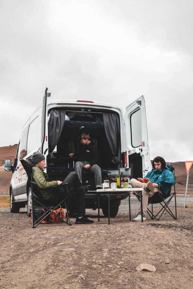 A group of people seated in chairs beside a parked campervan, engaged in conversation and enjoying their time together.