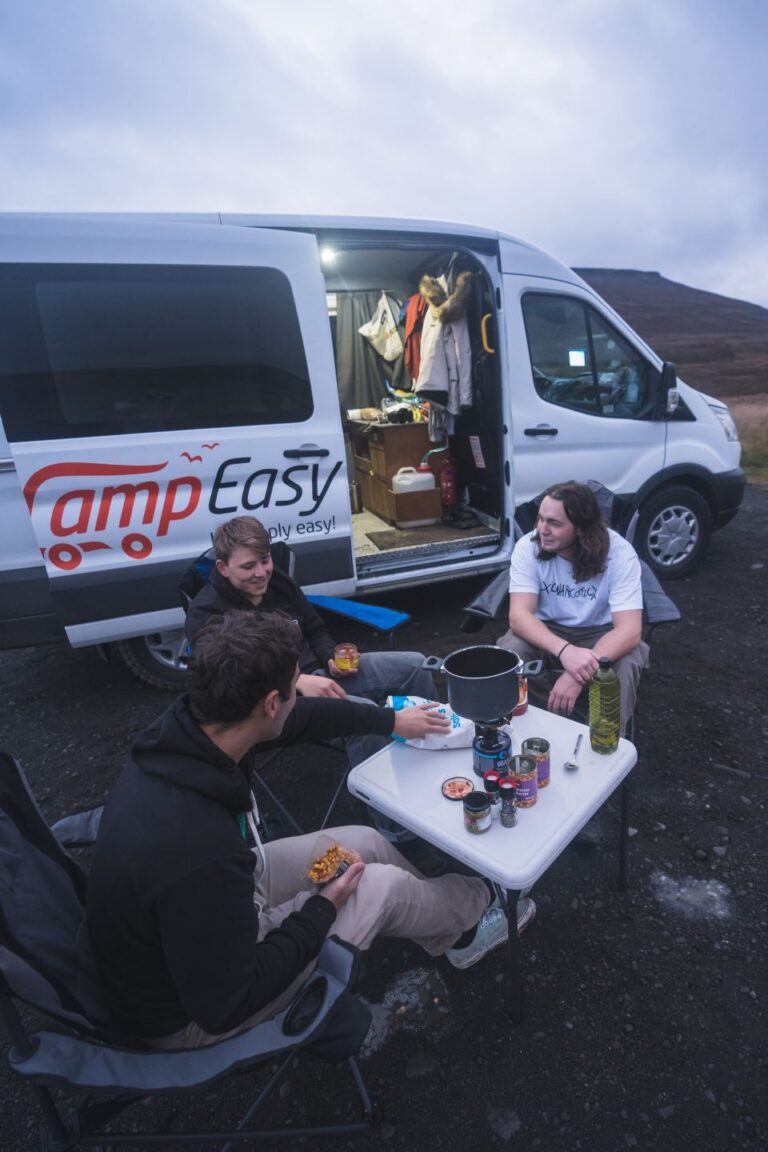 Three people sit around a portable table near a campervan, preparing a meal with a stove and various food items under a cloudy sky in Iceland.