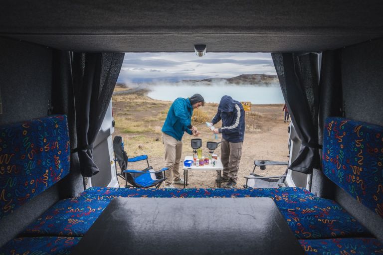 Two people are cooking a meal outside of the campervan near the steaming geothermal pool in the background.