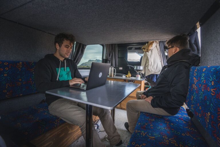 Two men are sitting inside the campervan. One is cooking a meal, and the other is working on his laptop.