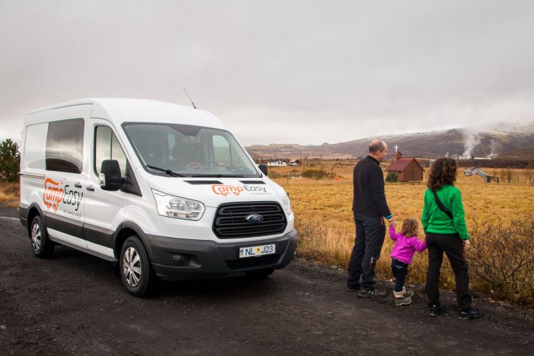 A family of three is watching the typical autumn view of the Icelandic countryside next to the parked white camper van.