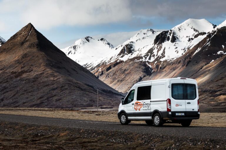 A white camping van parked on a dirt road surrounded by Icelandic partially covered by snow mountains.
