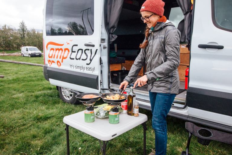A woman is preparing the food on the table next to the big white camper van.