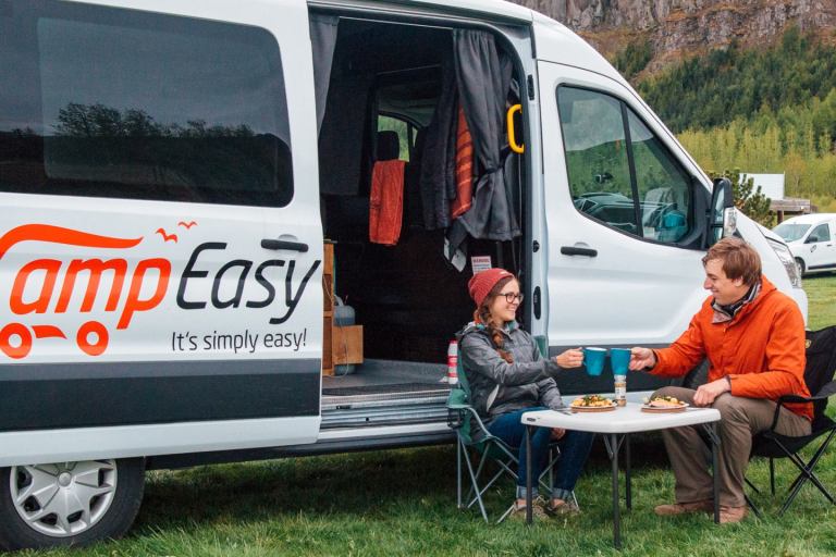 A couple is enjoying their meal while sitting on a camping chair next to the big white camper van parked on the green camping field.