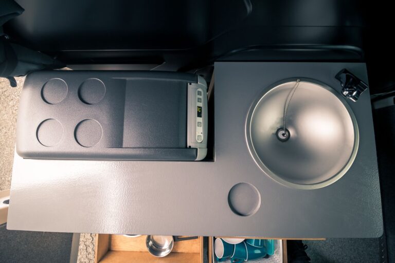 Interior view of a campervan kitchen corner, featuring a fridge, sink, and open shelves.
