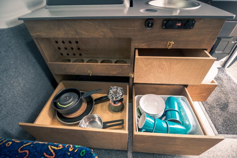 Interior view of a campervan kitchen corner, featuring a fridge, sink, and open shelves.
