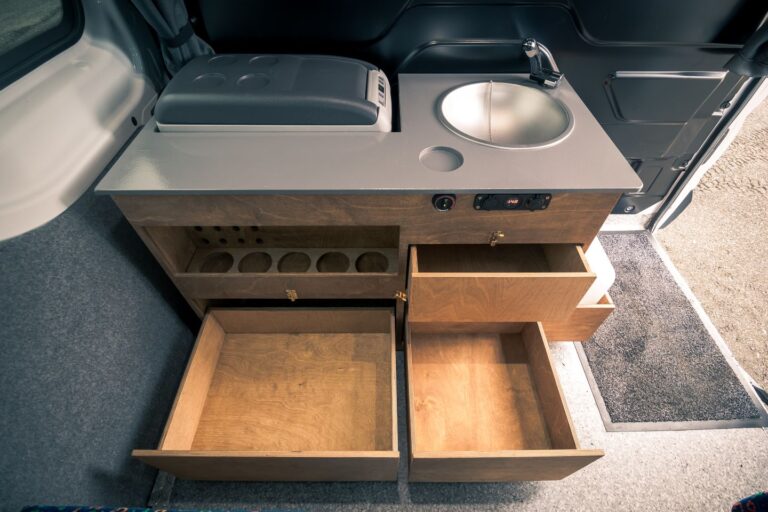 Interior view of a campervan kitchen corner, featuring a fridge, sink, and open shelves.
