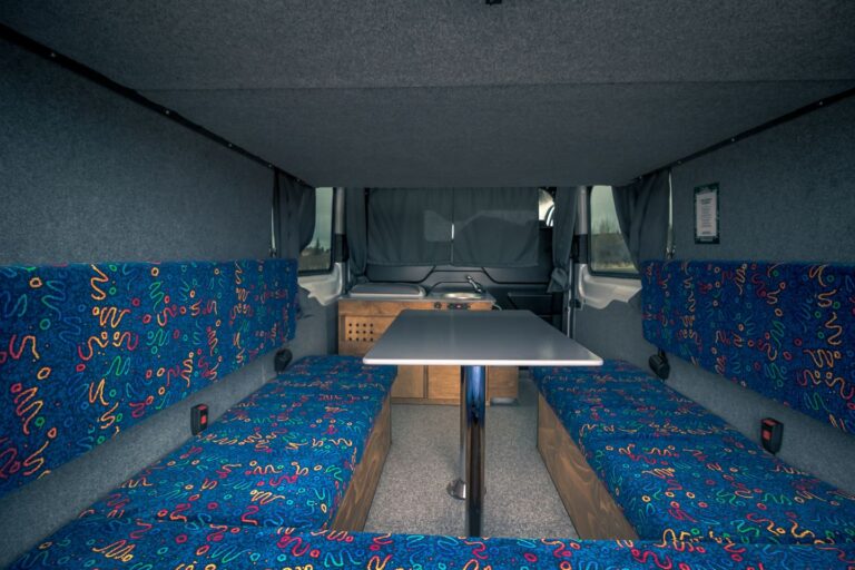 Interior view of a campervan with an assembled table, sitting area and kitchen corner in the background.