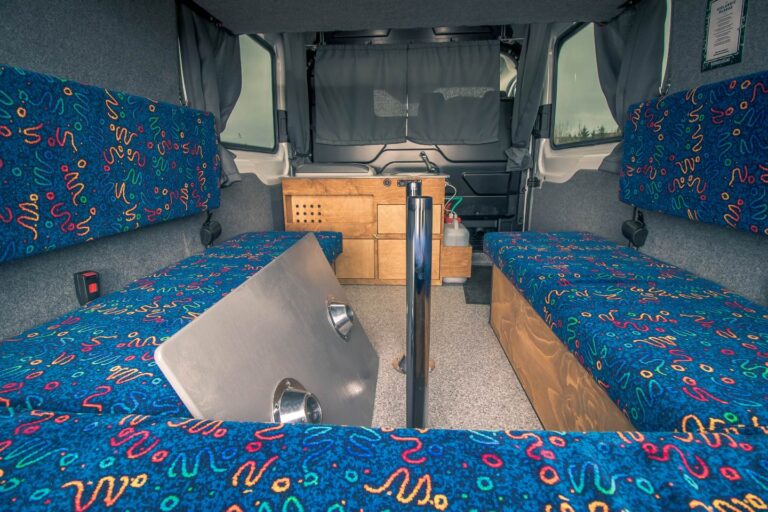 Interior view of a campervan with an unassembled table, sitting area and kitchen corner in the background.