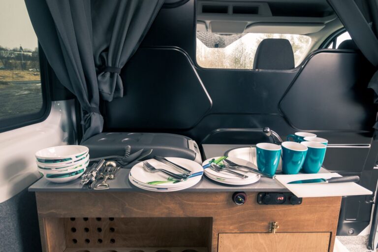 A kitchen corner inside a campervan showing set of cutlery, plates,cups, sink and fridge.