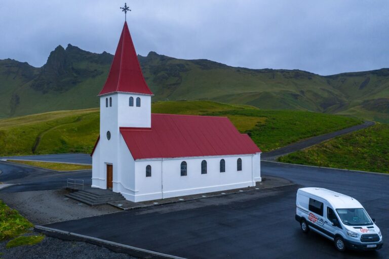 A white campervan parked beside a church in Iceland, surrounded by a scenic landscape.