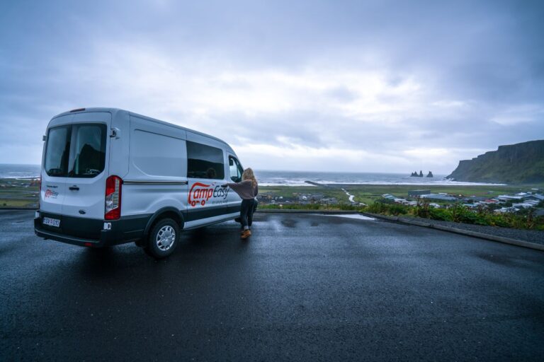 A woman stands beside a white campervan parked on gravel in Iceland, with clouds overhead and a road and the city of Vik in the background.