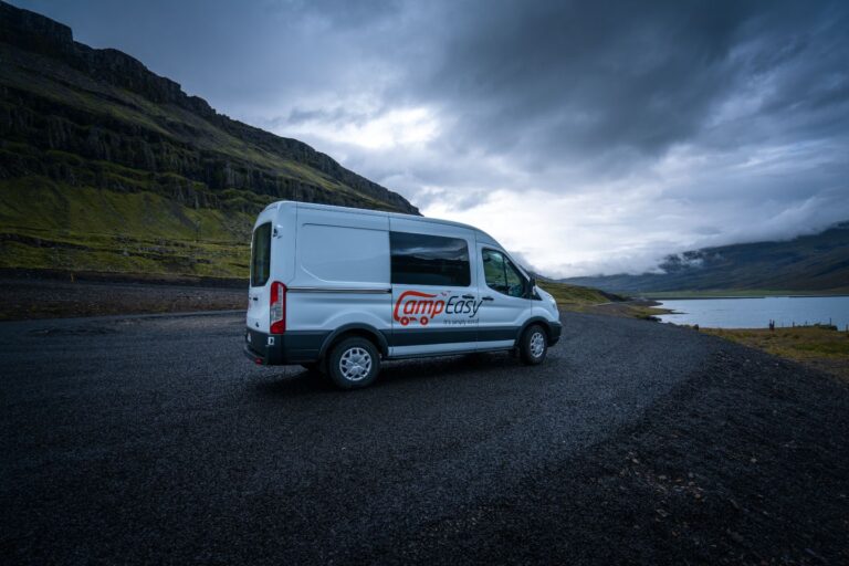 A campervan parked on gravel beside a road in Iceland, with mountains and a fjord in the background.