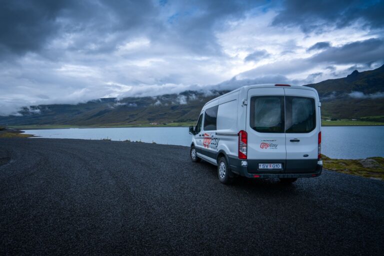 A campervan parked on gravel beside a road in Iceland, with mountains and a fjord in the background.