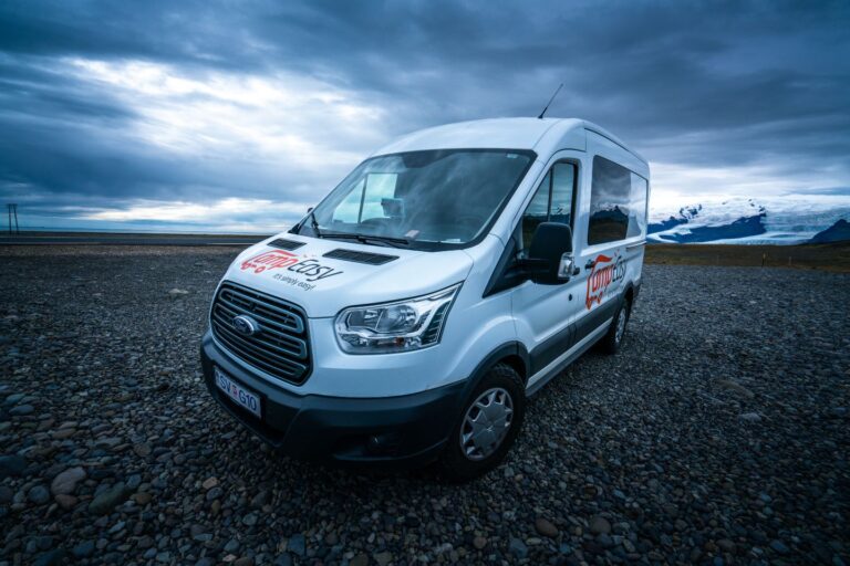 A white van on a gravel road under a dark sky, with mountains rising in the background, showcasing Iceland's landscape.