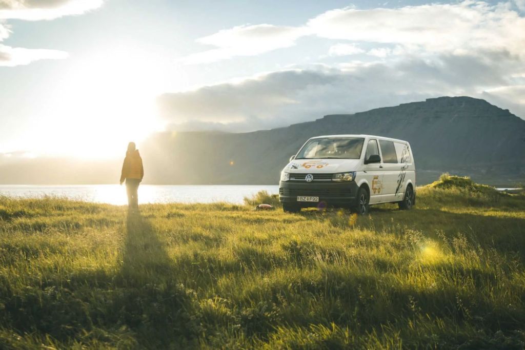 A person in a yellow jacket stands on a grassy field by a lake, with a white CampEasy camper van parked nearby. The sun is shining brightly over the mountains in the background, creating a serene and picturesque scene.