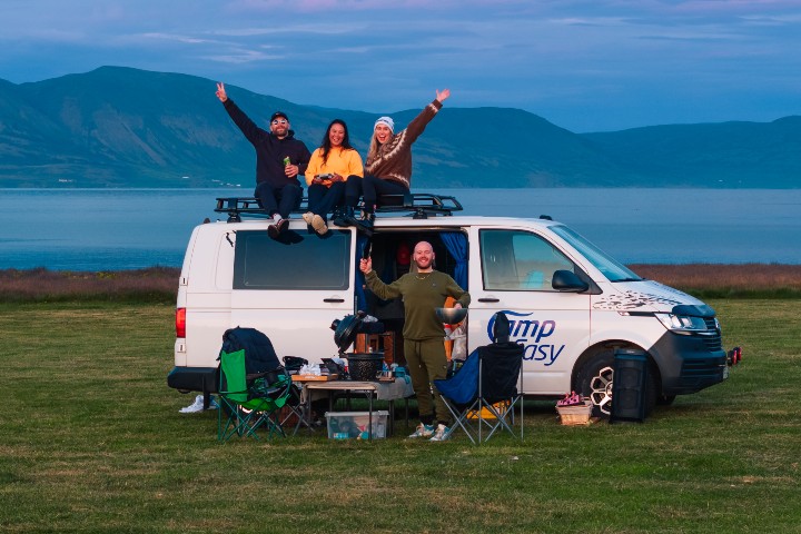 A group of friends enjoying a camping trip by the sea with a CampEasy camper van, set against a backdrop of mountains and ocean.