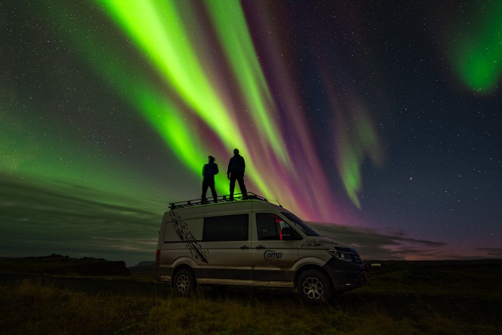 Two people standing on top of a CampEasy camper van under a vibrant display of the Northern Lights in Iceland.