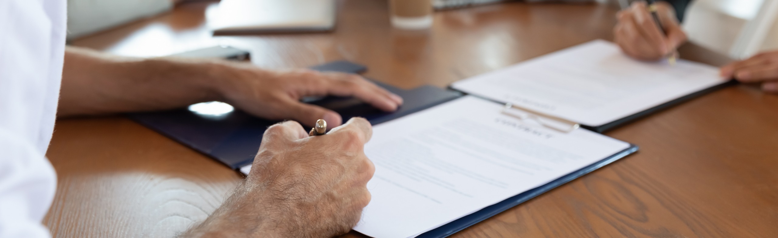 Close-up view of hands signing documents on a wooden table, focusing on the process of filling out forms or contracts.