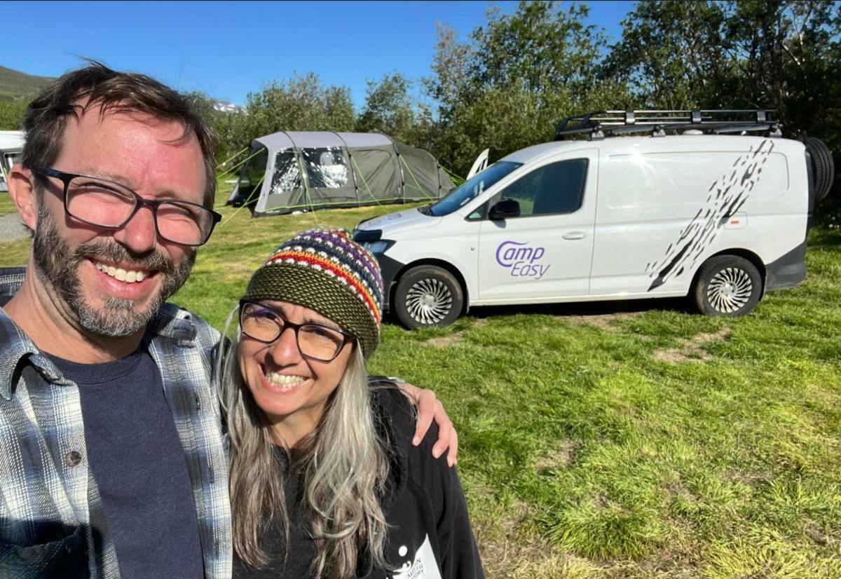 Happy couple taking a selfie with a CampEasy campervan in the background, parked at a vibrant camping site with tents and lush greenery, showcasing a joyful travel experience in Iceland.