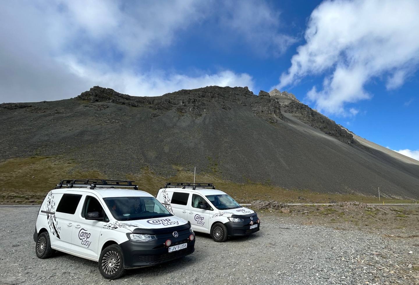 Two white CampEasy campervans parked on a rocky terrain with a large mountain looming in the background under a bright blue sky, perfect for group expeditions in Iceland's dramatic landscapes.
