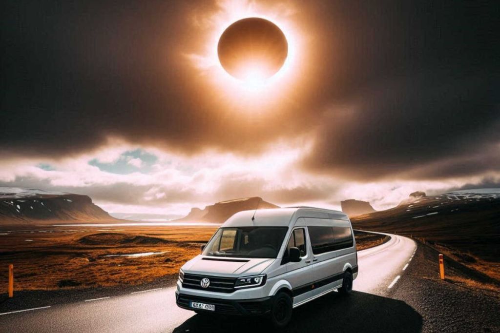 A white camper van is parked on a scenic road in Iceland, surrounded by dramatic landscapes with mountains and fields. Above, a solar eclipse is visible in the sky, casting a dark shadow over the land and creating a striking, ethereal atmosphere. The road stretches into the distance, emphasizing the sense of adventure and the unique experience of viewing the eclipse from this remote location.