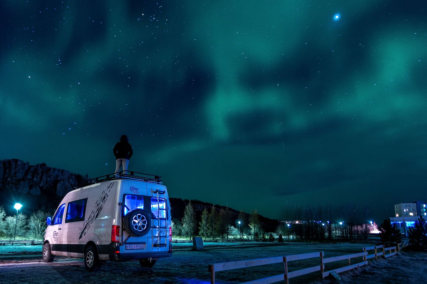 Campervan parked under the northern lights in Iceland, with a person standing on the roof admiring the glowing night sky.