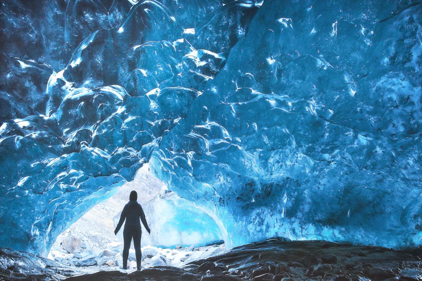 A person stands in awe inside a glowing blue ice cave, surrounded by smooth, translucent frozen walls.
