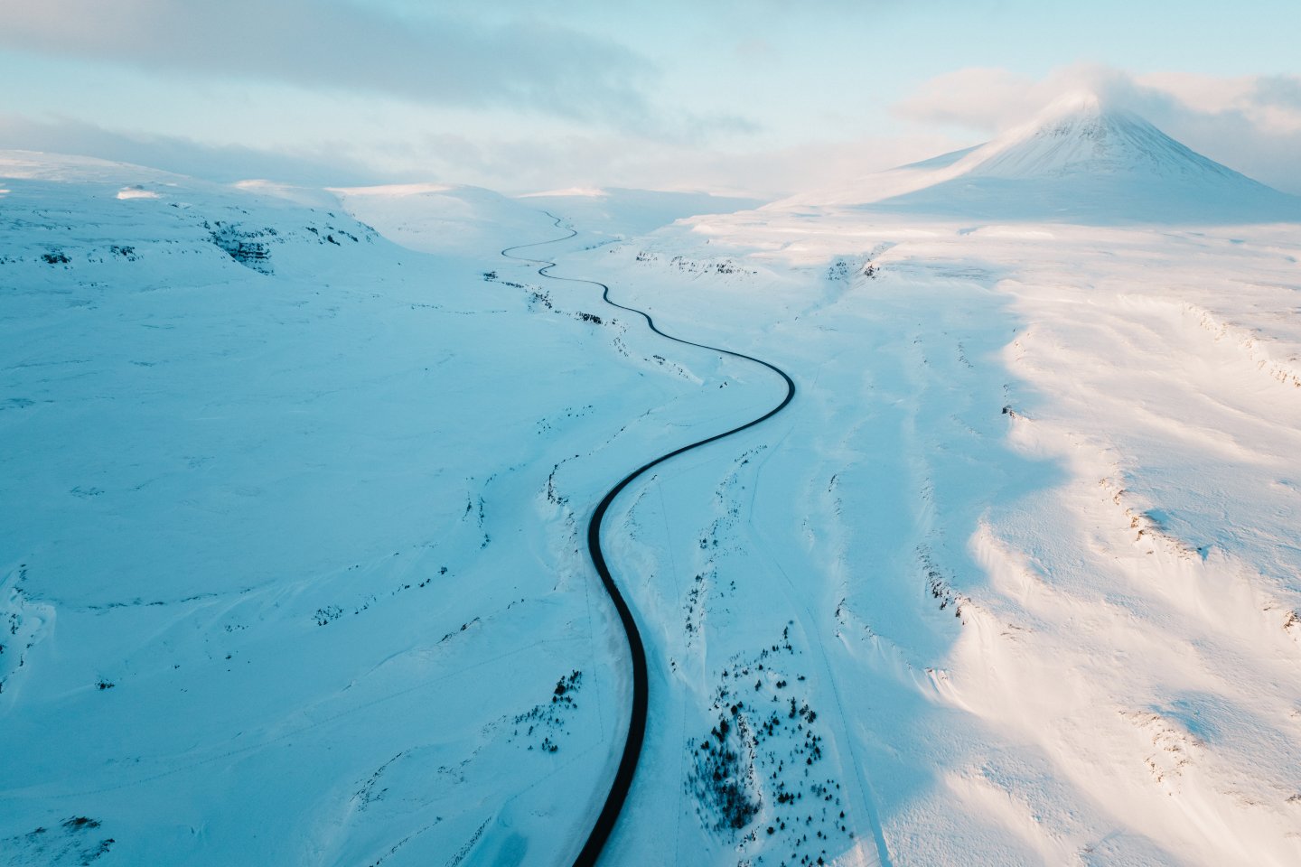 A winding road cuts through a vast, snow-covered Icelandic landscape, with a towering mountain partially covered in clouds.