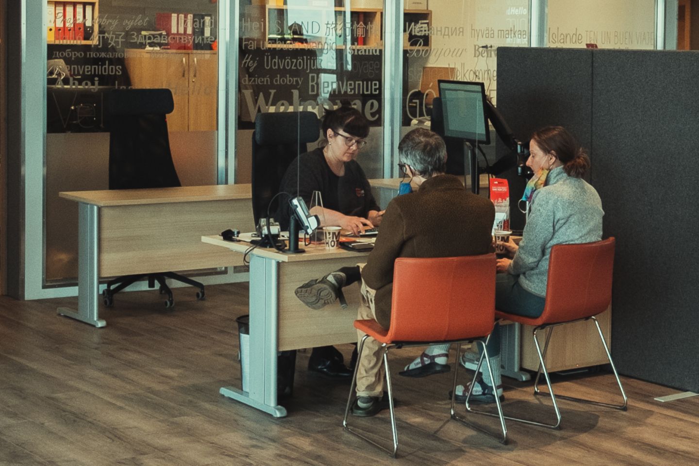 A CampEasy staff member assisting two customers at a desk in a modern office, with walls decorated with welcome messages in various languages.