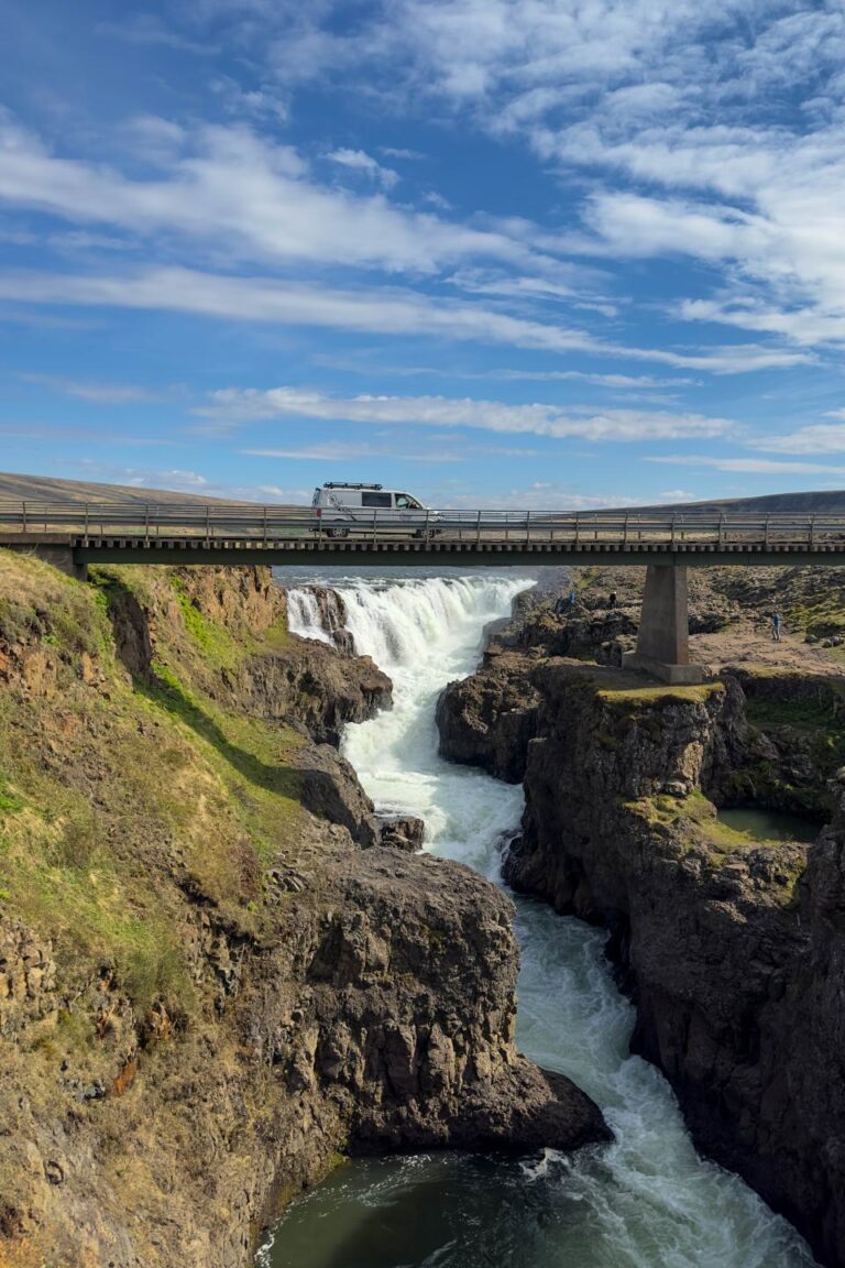 A campervan driving across a bridge next to a spectacular waterfall in Iceland.
