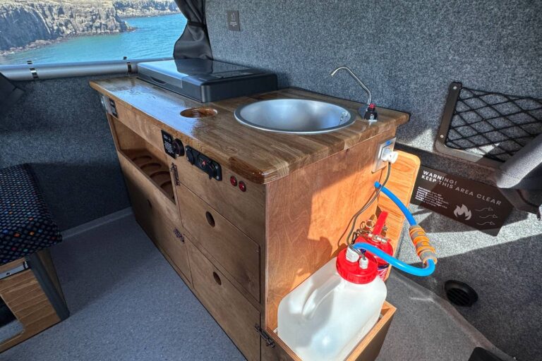A kitchenette inside the campervan, including a sink, fridge, water tank and shelves with cabinets.
