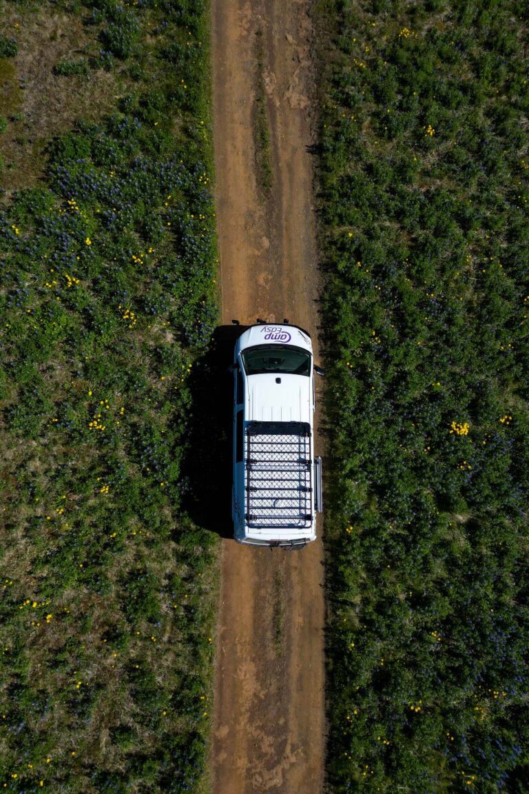 A bird's-eye view of a campervan parked on a sandy road surrounded by green fields with yellow flowers.