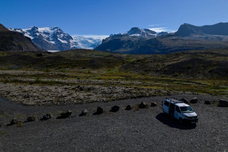 A camper parked on a gravel parking lot next to green hills, mountains, and a glacier visible far in the distance.