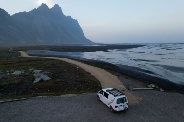 A camper parked on a gravel parking lot next to a beach with black sand and huge mountains.