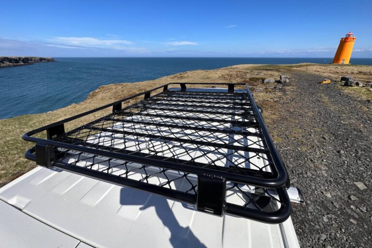 The photo focuses on the roof rack of a camper parked next to a bluff by the ocean and an orange lighthouse.