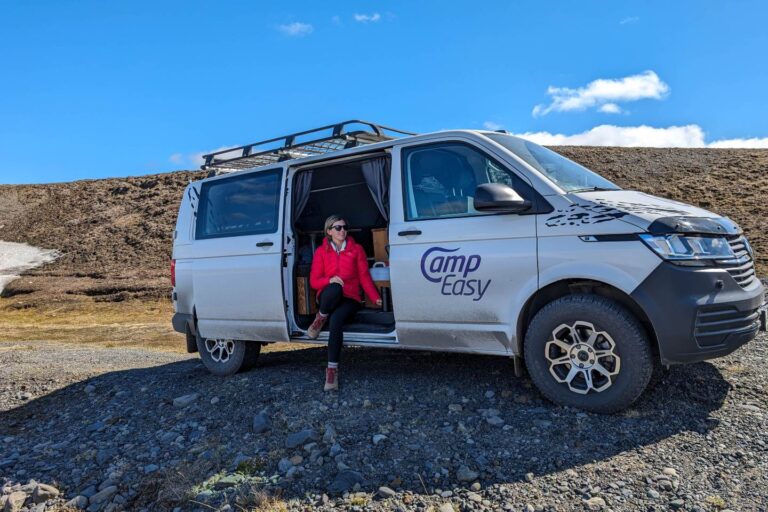 A woman in a red jacket sits on the floor of a campervan in the open side door of a camper parked on the gravel next to a hill.