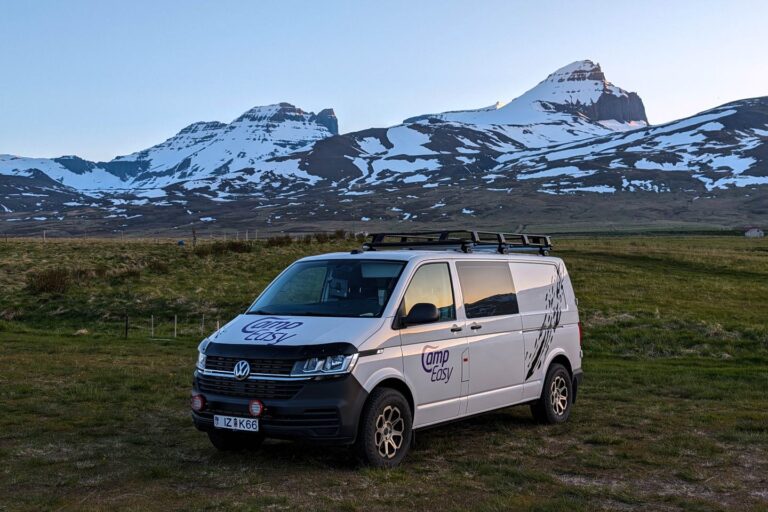 A campervan parked on the grass with fields of grass and partially snow-covered mountains behind it.