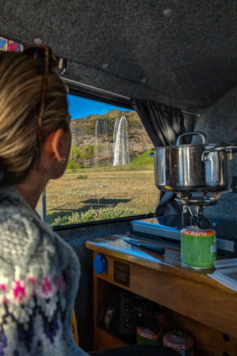 A woman cooking in a pot inside a campervan looks out the side window at a nearby waterfall.
