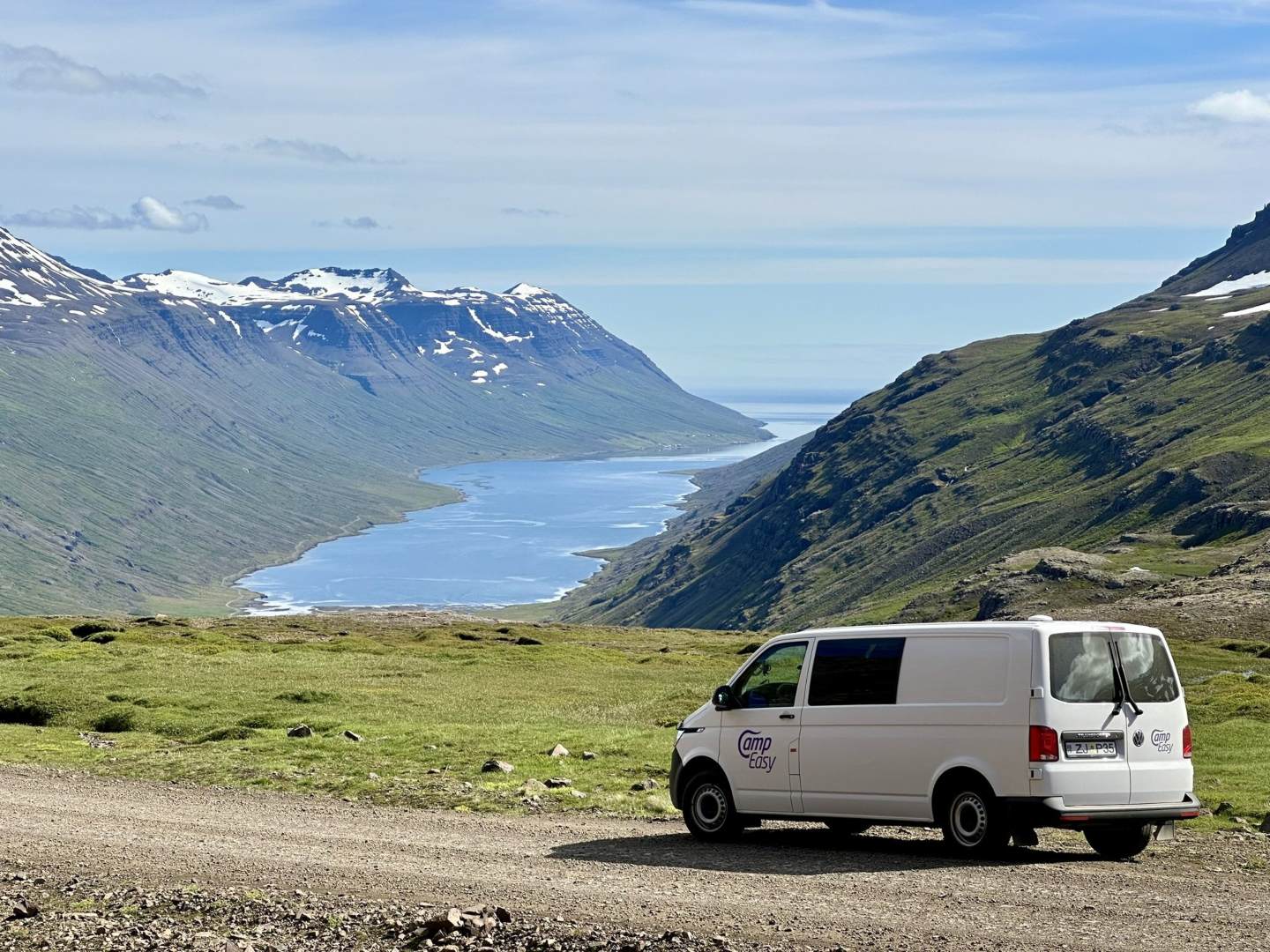 CampEasy campervan parked on a gravel road with a panoramic view of a fjord flanked by majestic mountains, showcasing Iceland's stunning landscapes perfect for exploratory travels.