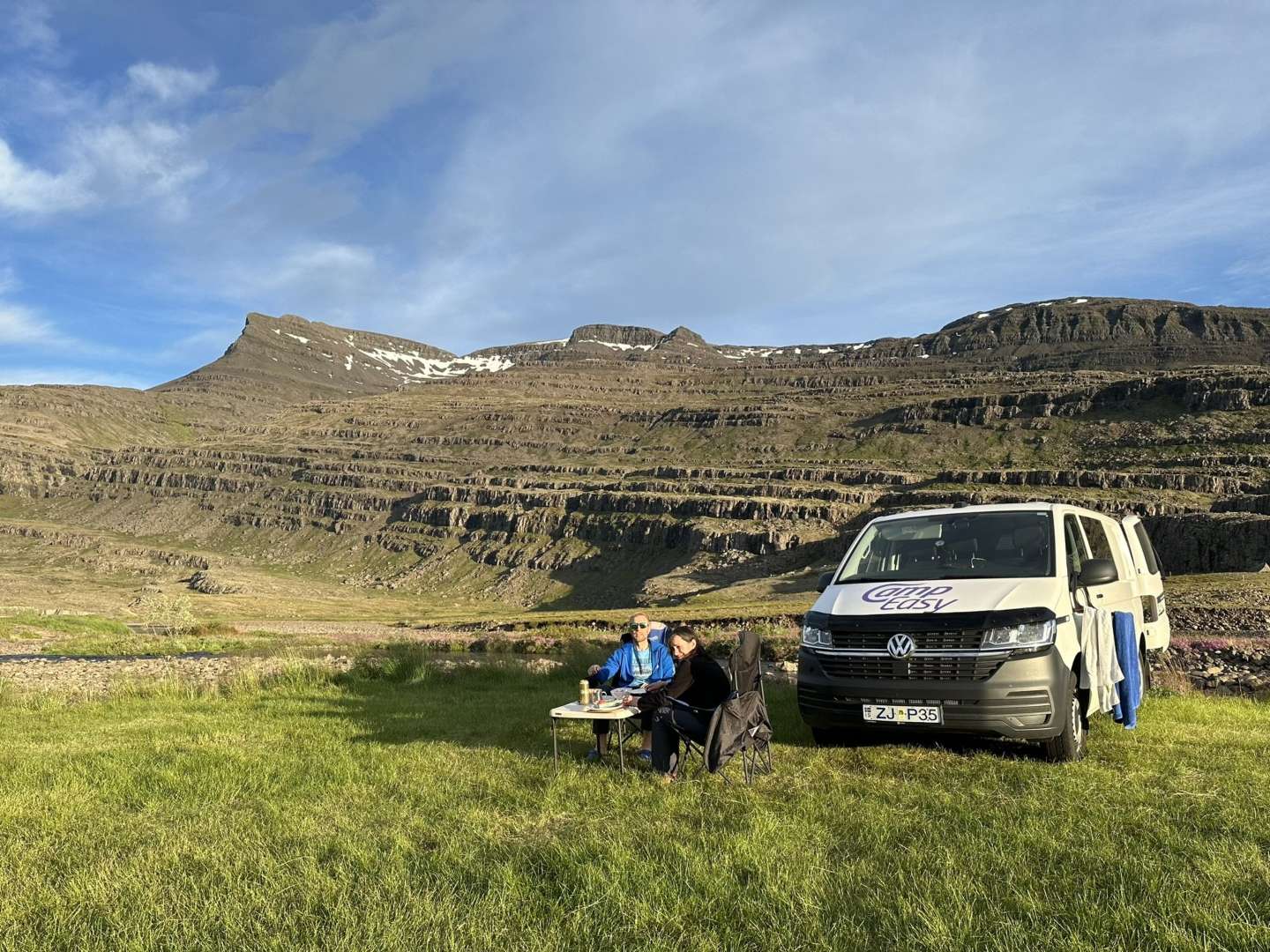 Two people relaxing beside a CampEasy campervan with a picnic table, set against the backdrop of a layered mountain in Iceland, enjoying the peaceful outdoor environment.