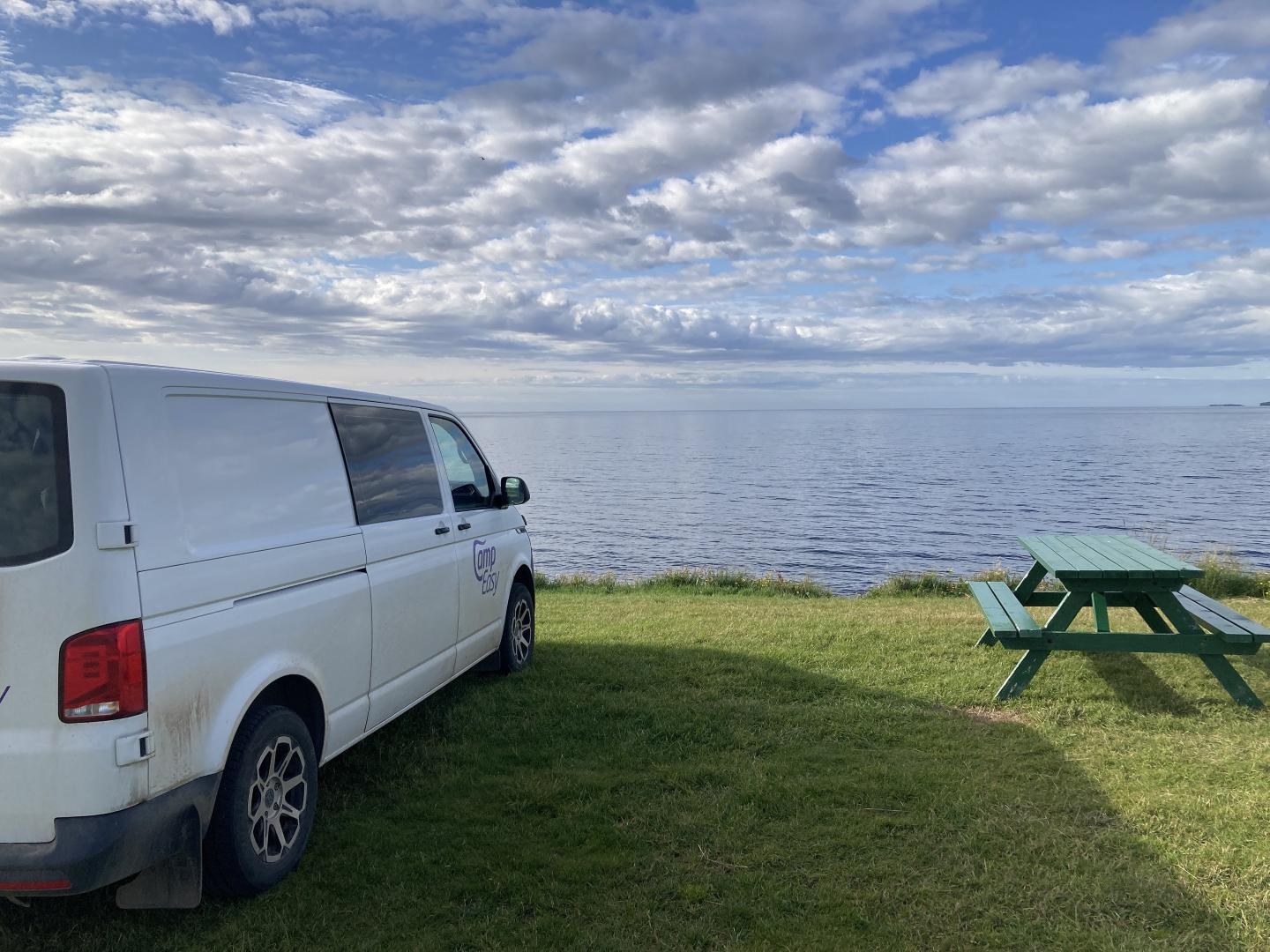 CampEasy campervan parked beside a calm sea with a green picnic table on the grass, under a sky dotted with clouds, offering a serene setting for coastal relaxation in Iceland.