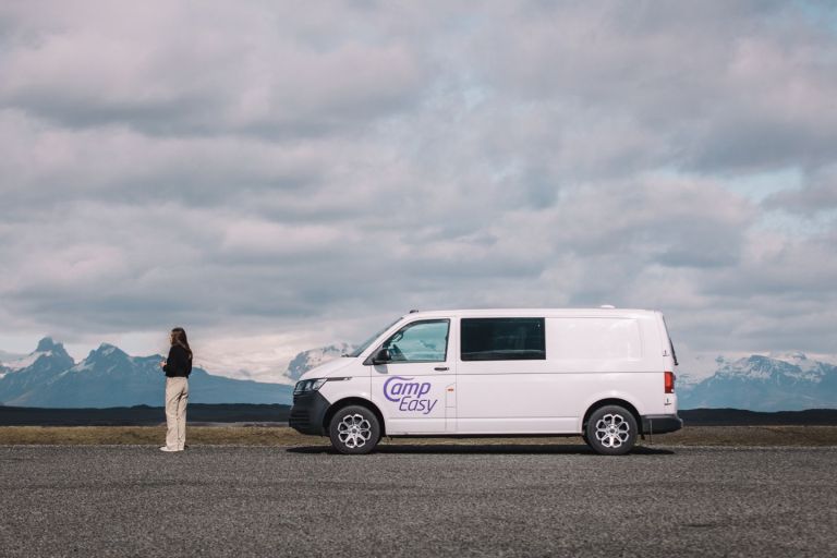 Woman standing in front of white campervan parked on the road with the snowy mountains in the backgorund.