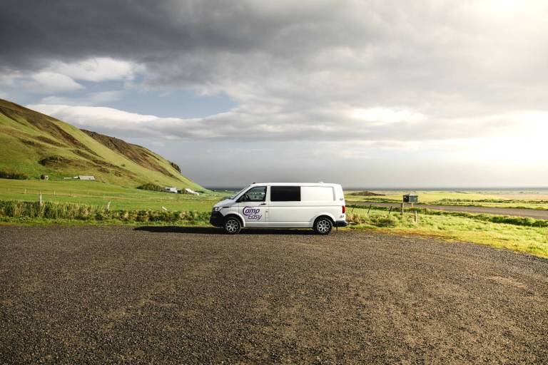 White Camper van parked on the gravel parking next to the green fields and mossy mountains.