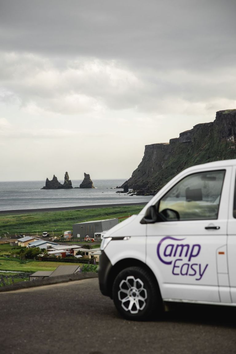 A white camper van parked on the parking with view on the buildings, grassy field, black sand beach and famous rock formation and cliffs in Iceland