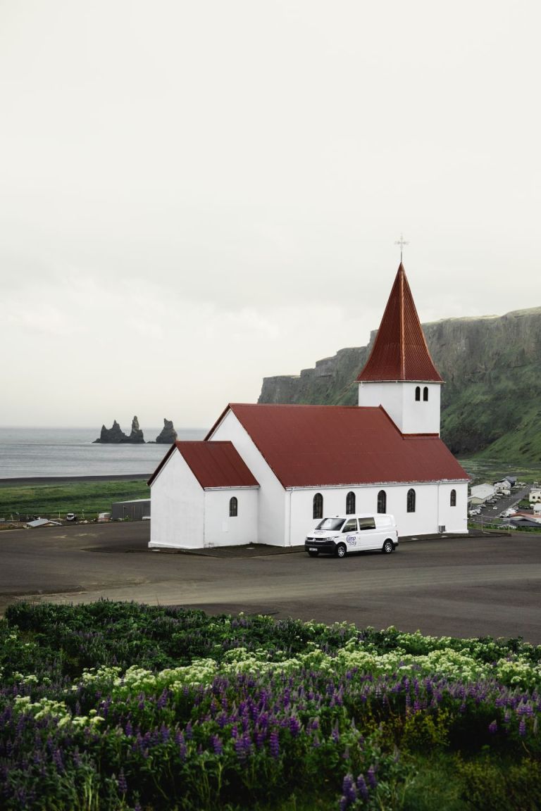 White church with a red roof and a 'Camp Easy' van parked nearby, with ocean and rock formations in the background.