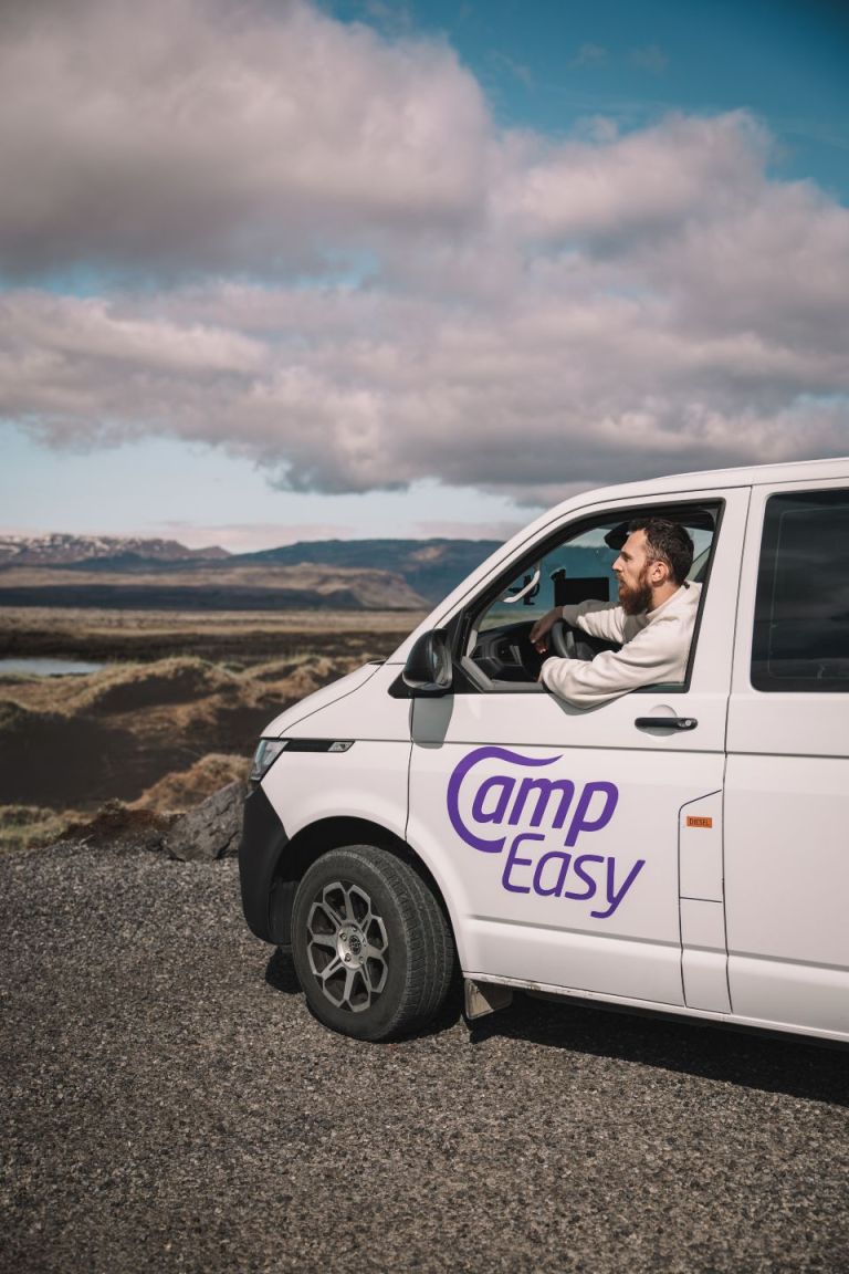 A man looks out the open window of a white camper van parked on a rocky parking lot. There are mountains and clouds in the background.