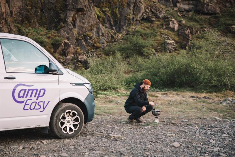Man cooking near the white camper van parked in the rocky parking. In the background there are typical Icelandic mossy cliffs and rocky terrain.